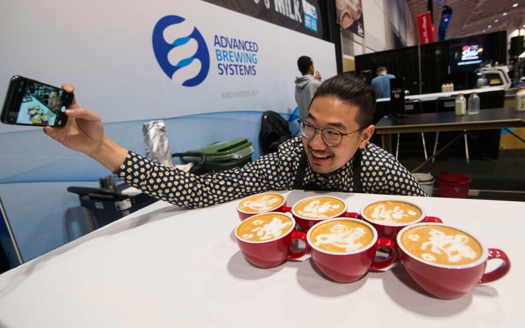 A barista takes a selfie with latte art on six milk-based drinks.