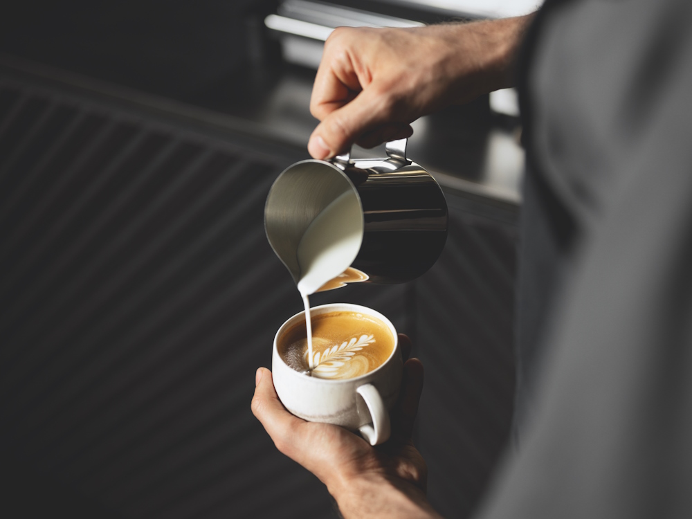 A barista pours latte art in a white ceramic cup.